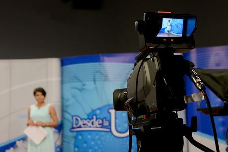 En primer plano se aprecia una cámara de televisión y al fondo la presentadora frente al rótulo del programa Desde la U, del Canal UCR.