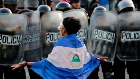 Imagen de una persona con una bandera de Nicaragua frente a un grupo de policías que están obstruyendo el paso.