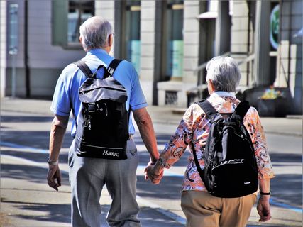 Imagen de espalda de un hombre y una mujer, adultos mayores, tomados de la mano caminando por una plazoleta.